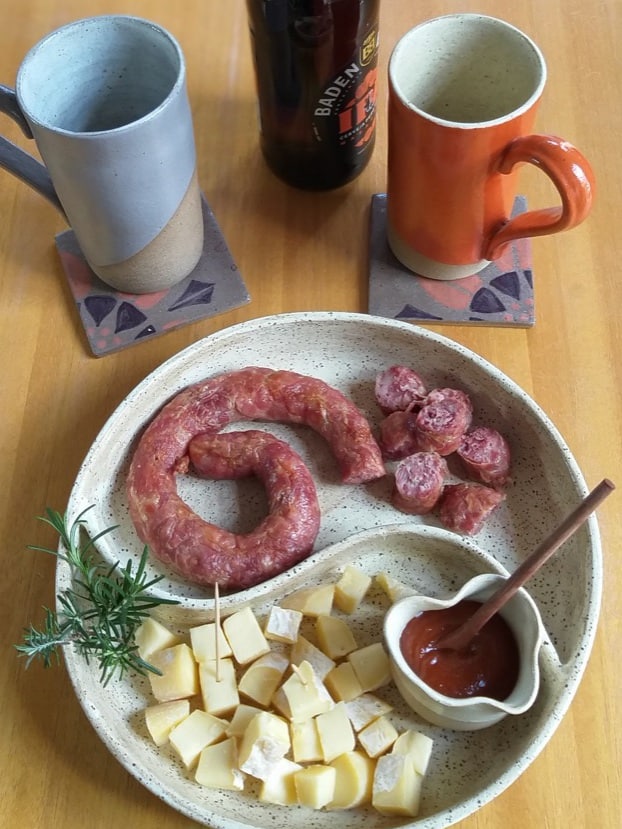 Foto feita de cima mostra uma petisqueira bege, com divisórioa em forma de S, contendo linguiça frita e cubos de queijo. Ao fundo, há duas canecas de cerâmica: uma cinza e uma laranja.