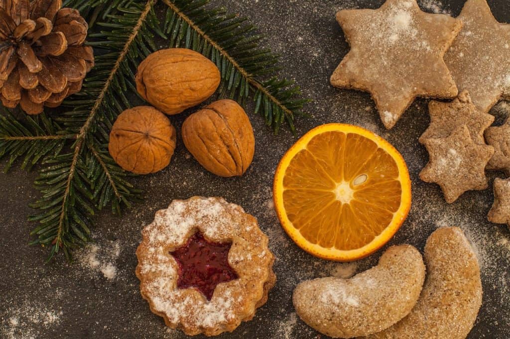 Sobre uma superfície marrom, vemos biscoitos, nozes com casca, uma pinha, um galho de pinheiro, e uma rodela de laranja. 