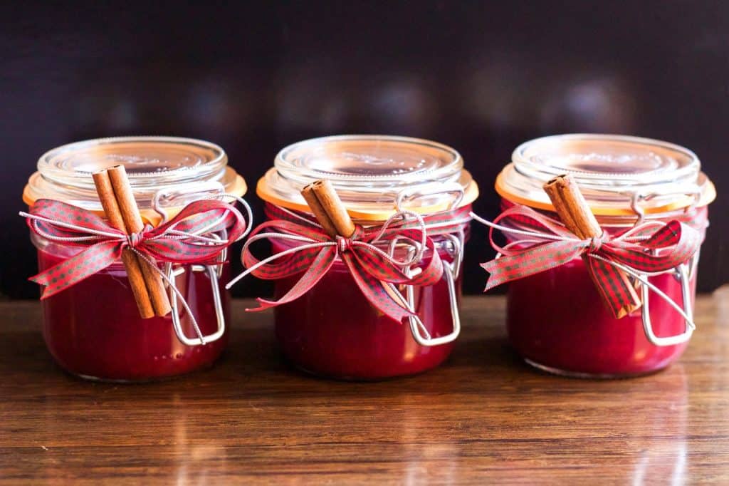 Três potes de vidro, herméticos, contendo geléia vermelha. Há um pedaço de canela em pau amarrado aos vidros com fita xadrez de vermelho e verde. 