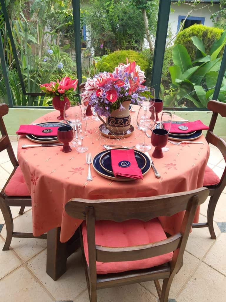 Mesa de Natal decorada com  4 lugares. A toalha e os guardanapos são  vermelhos. Os pratos são de cerâmica azul marinho. No centro há uma jarra com arranjo de flores azuis e vermelhas. 