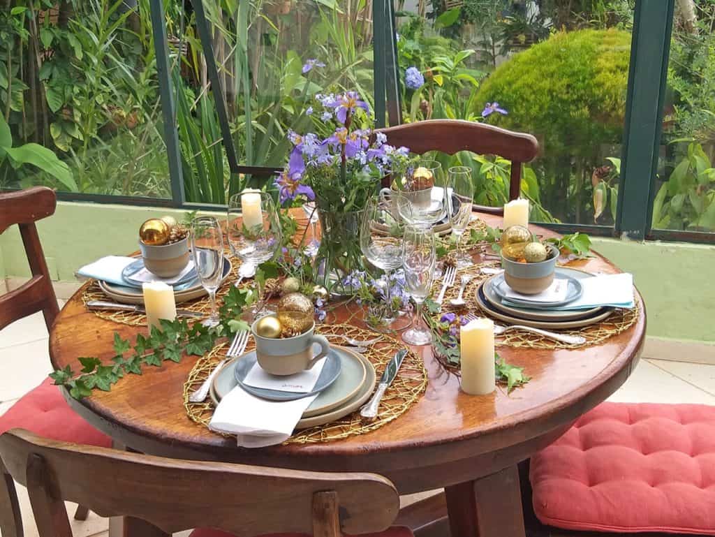 Mesa de Natal decorada, posta para 4 pessoas. Há xícaras e pratos de cerâmica azul claro. Dentro das xícaras, vemos bolas de Natal em tons de dourado. No centro da mesa ha um vaso com flores azuis. 