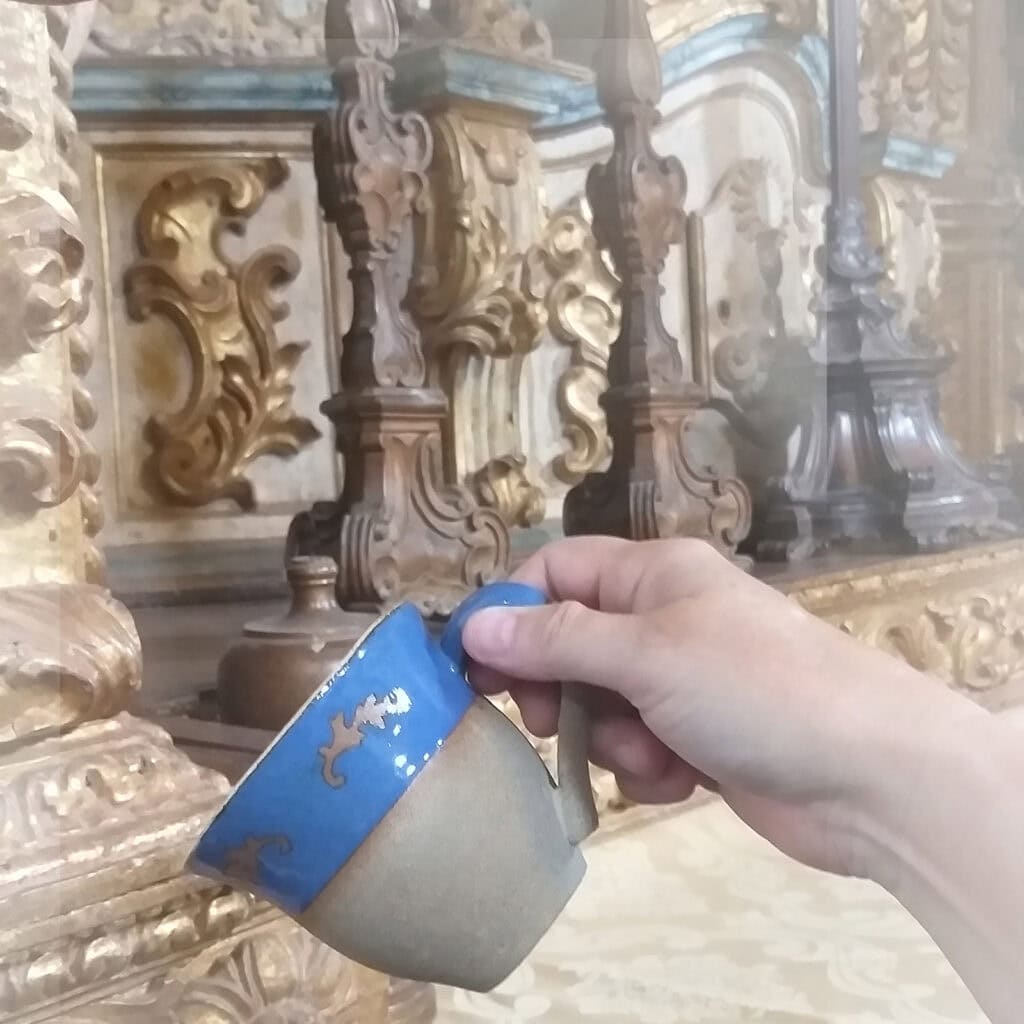 Mão de mulher segura uma xícara de cerâmica com faixa azul perto da borda e desenho de ramo de canto. Ao fundo, parte de uma altar barroca numa igreja.