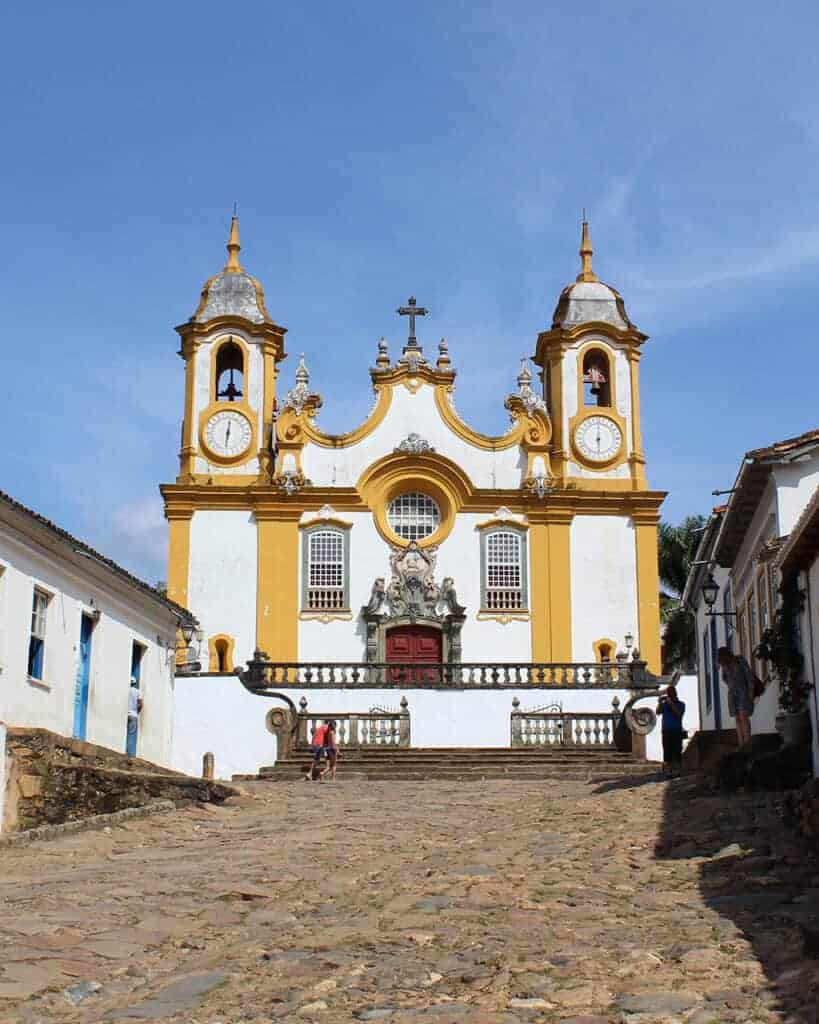 Foto colorida da fachada da igreja Matriz de Santo Antônio, em Tiradentes, MG. Ela tem duas torres com sinos, detalhes rebuscados típicos do Barroco brasileiro, e está pintada de branco com colunas e frisos em amarelo ocre. 