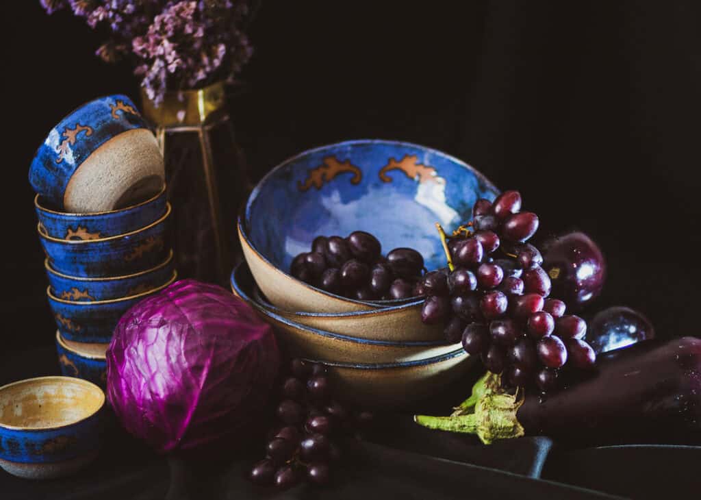 Num fundo preto, vemos tigelas empilhadas, cachos de uvas escurs e um repolho roxo.